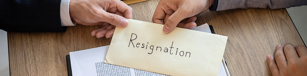A man hands his boss an envelope that reads 'resignation', across a wooden desk. Resignation Letter from Employment Law Friend. 